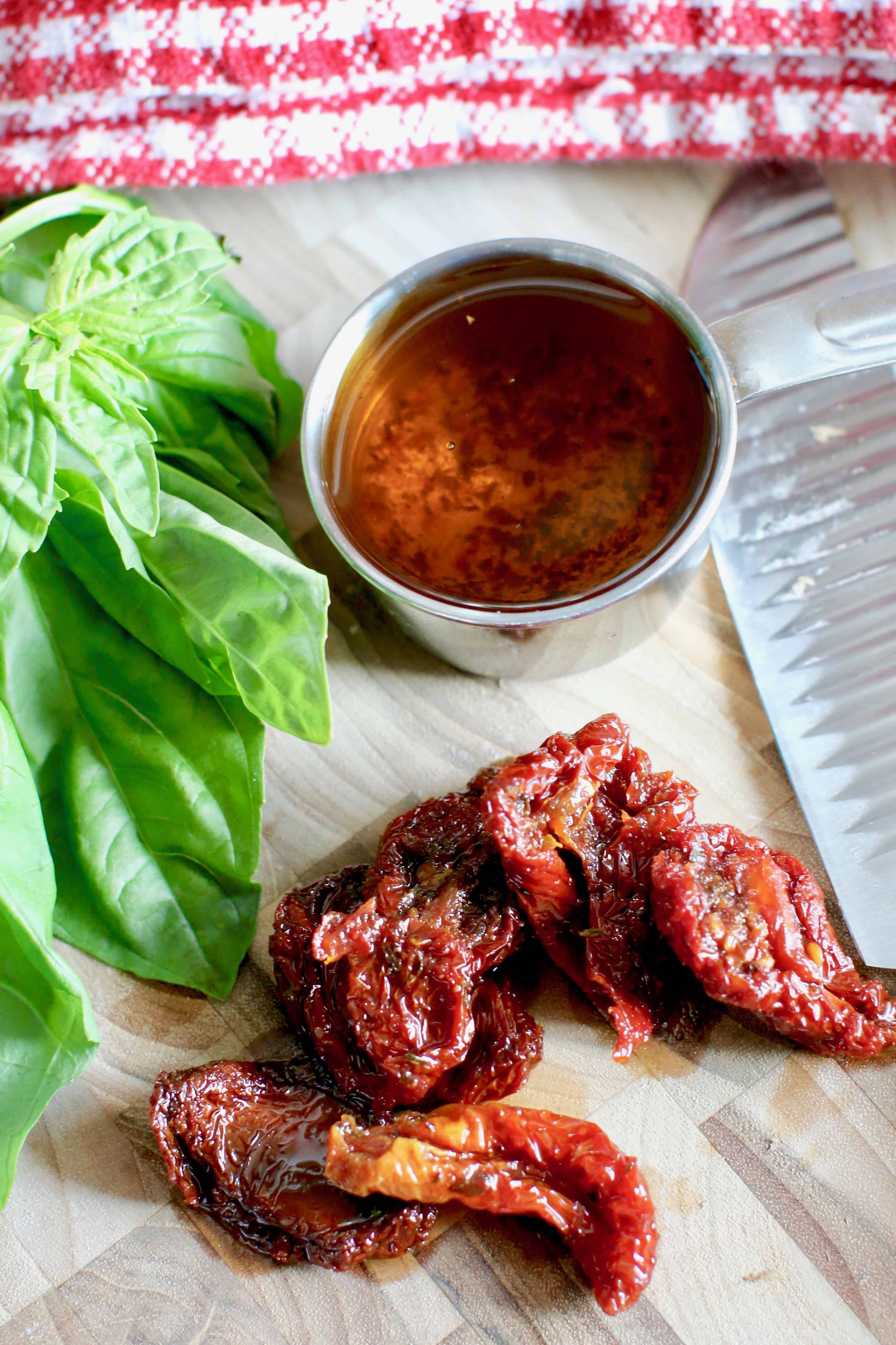 sun dried tomato oil shown in a measuring cup with several sun-dried tomatoes on a cutting board with fresh basil.