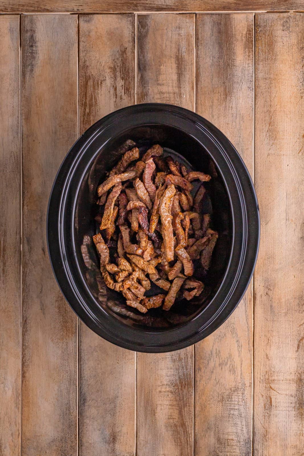browned beef strips shown in a black, oval slow cooker. 