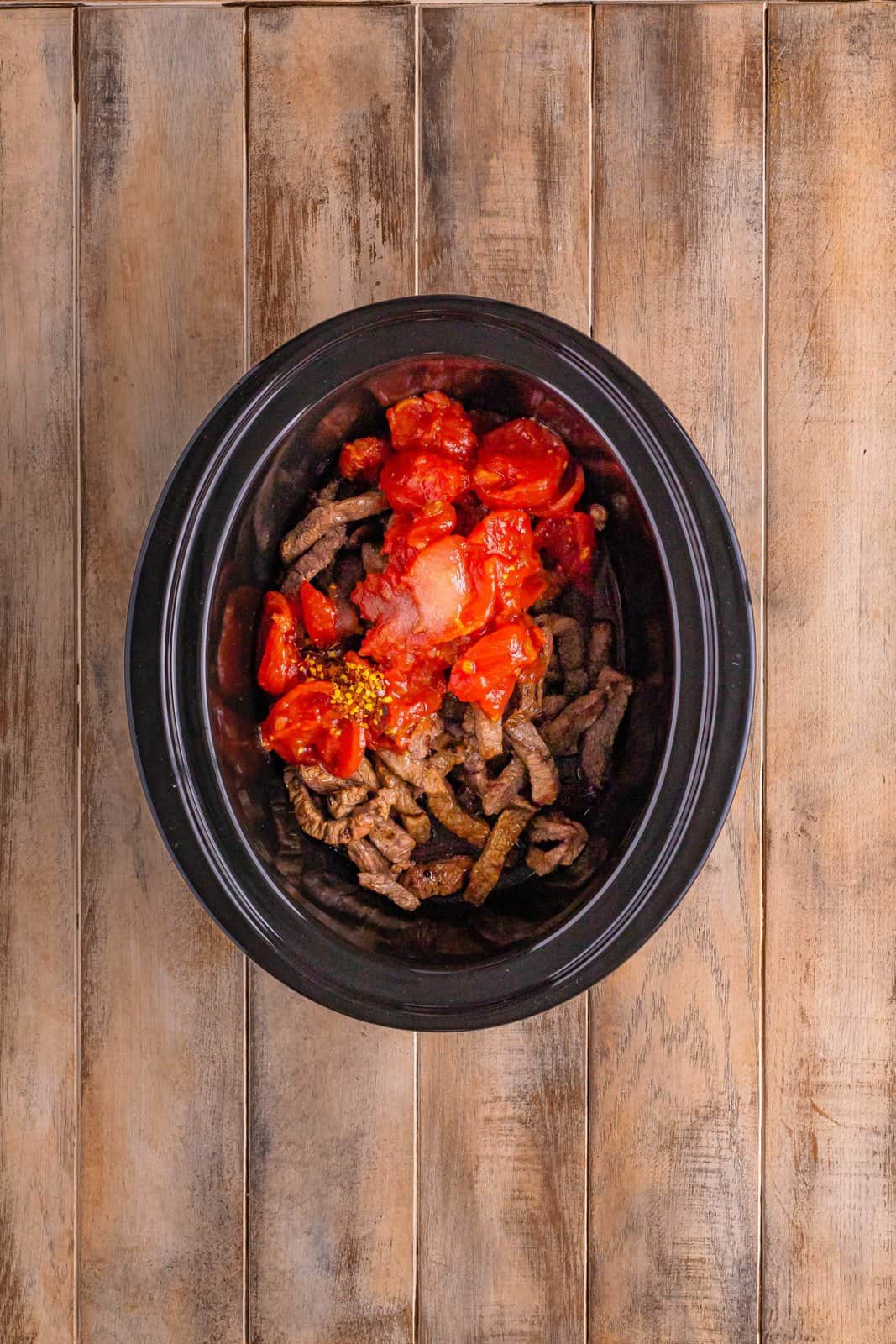 stewed tomatoes and red pepper flakes added on top of the pepper steak in the black oval slow cooker. 
