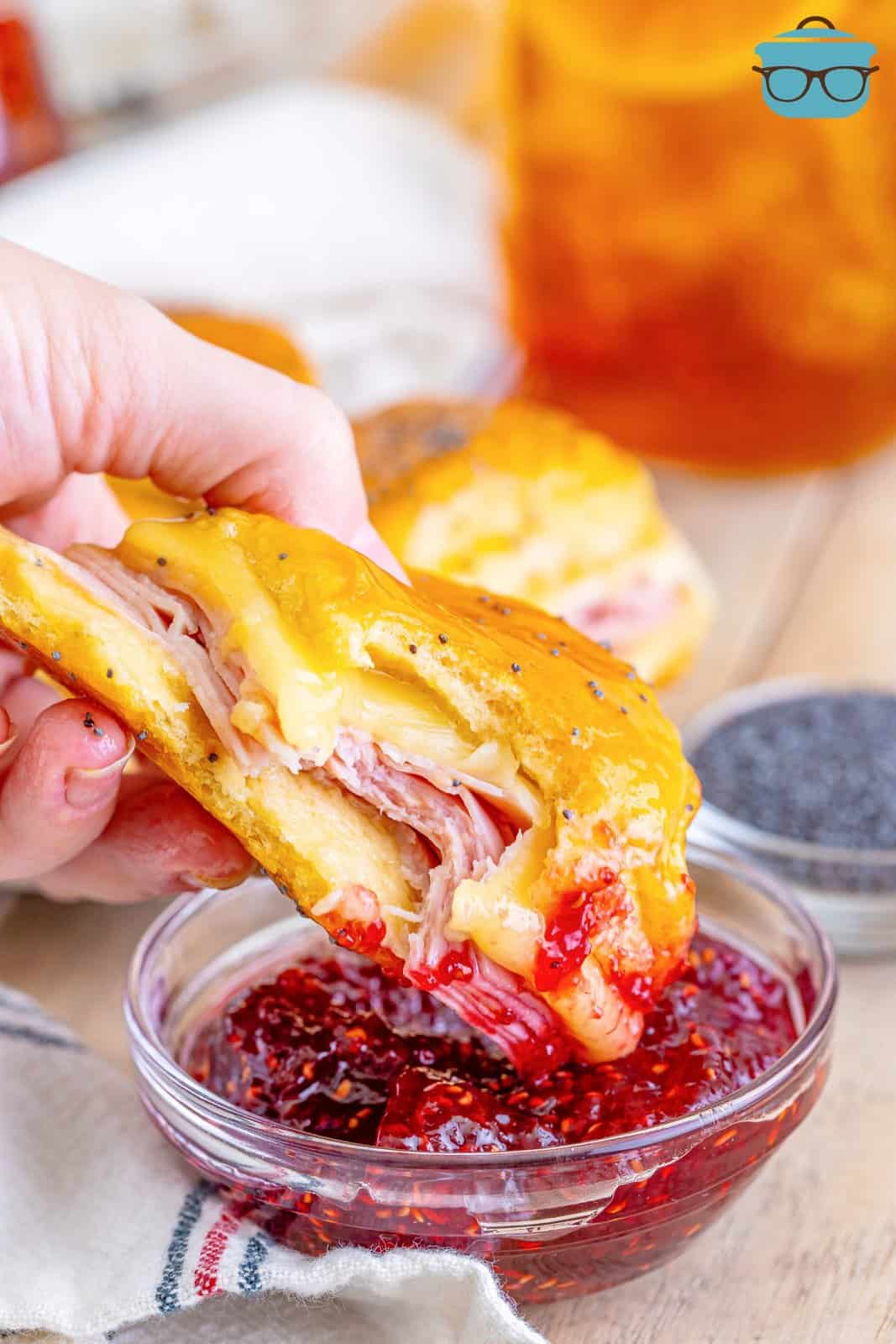 a hand dipping a glazed ham and cheese sliders into a bowl of raspberry jam.