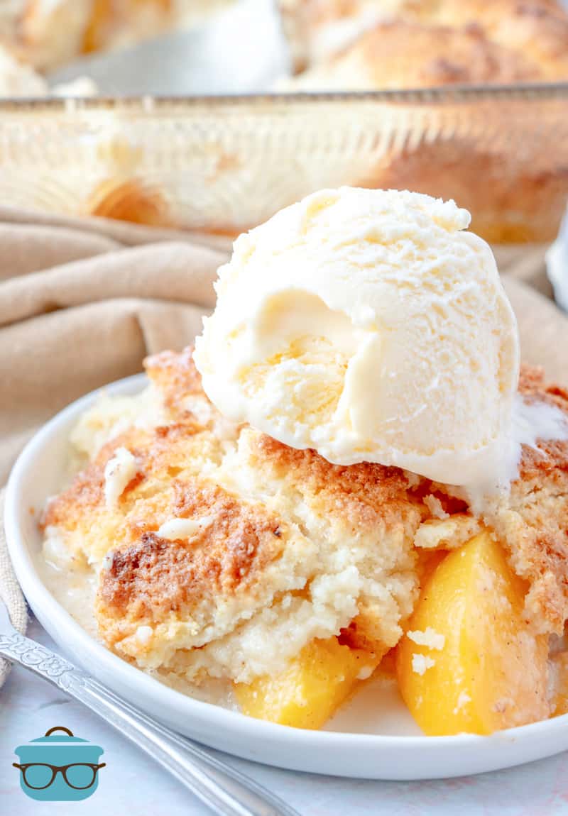 peach cobbler shown on a small round white plate with a scoop of vanilla ice cream on top.
