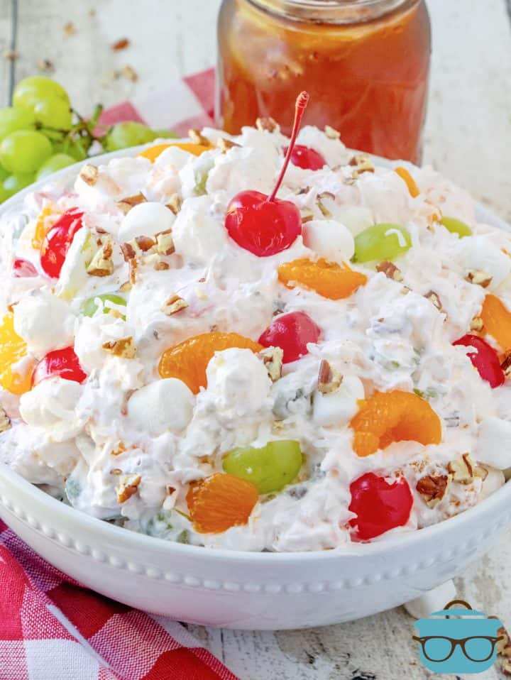 a large white bowl with ambrosia salad in it with a glass of iced tea in the background.