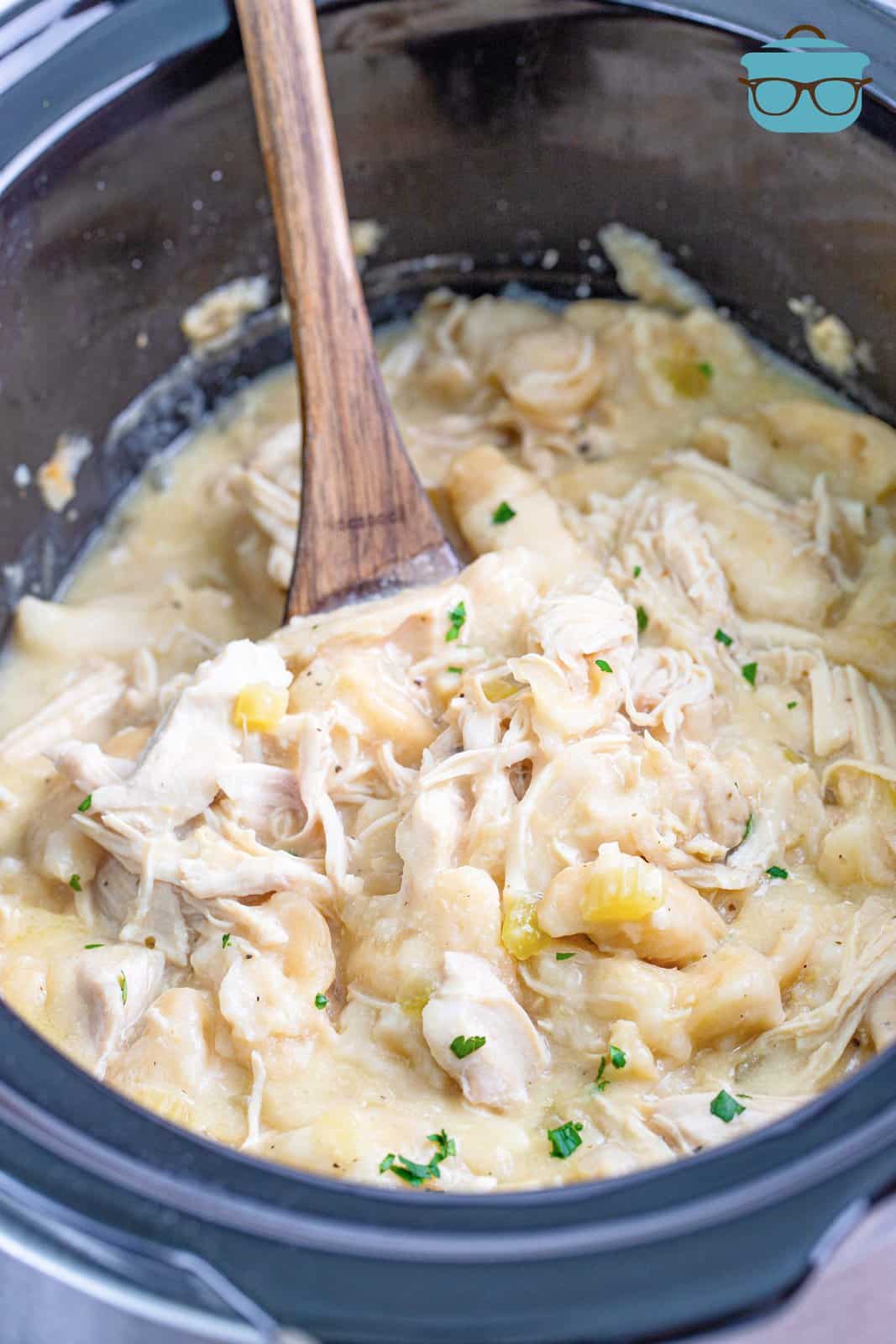 chicken and dumplings shown in a slow cooker with a wooden spoon. 