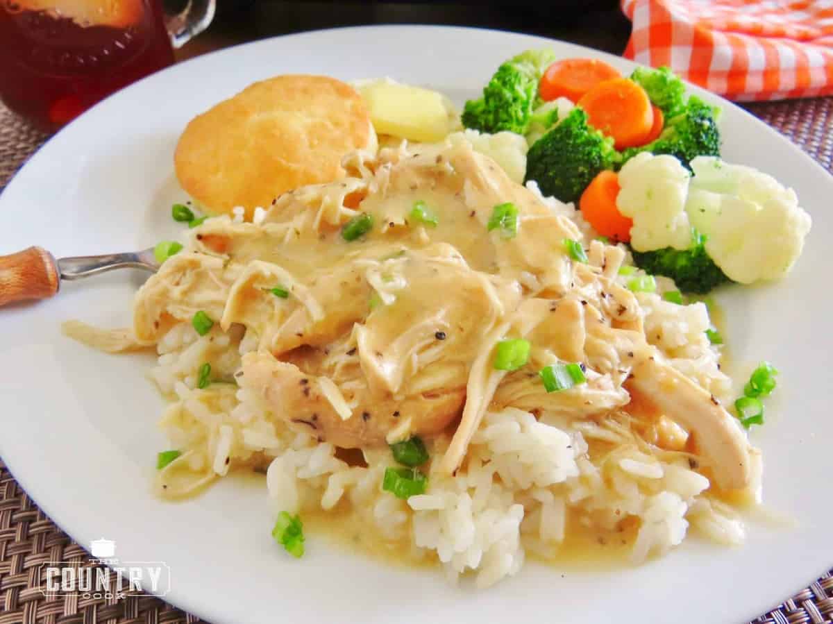 chicken and gravy served over rice on a white place with roasted vegetables and a biscuit on the sides.