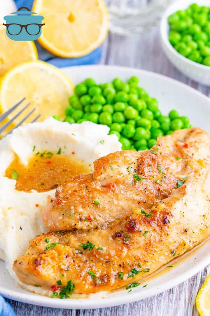 two slices of lemon garlic chicken tenders on a white plate with a side of mashed potatoes and peas.