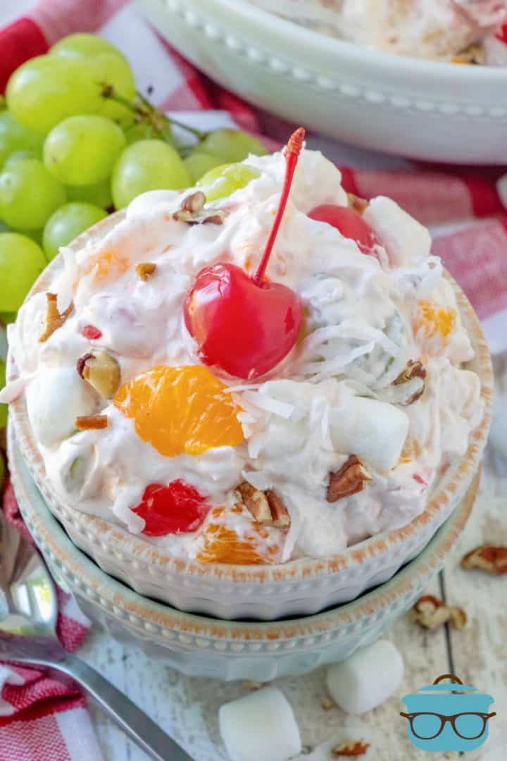 a serving of ambrosia fruit salad shown in a small white bowl with a maraschino cherry on top with a bunch of green grapes on the side.