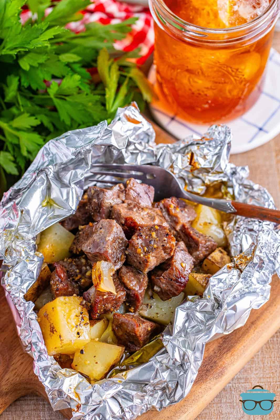 an overhead view of cooked steak and potato bites in aluminum foil packet with fresh parsley in the background.