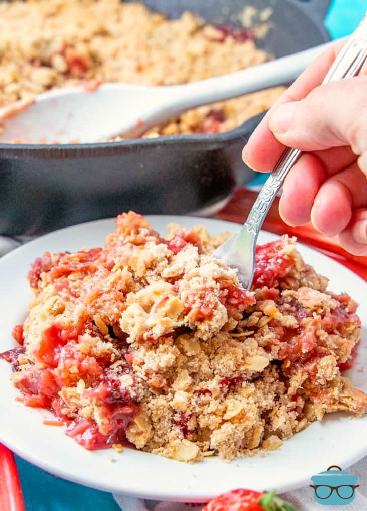 a fork being inserted into a serving of strawberry rhubarb cobbler that is on a white dessert plate. 