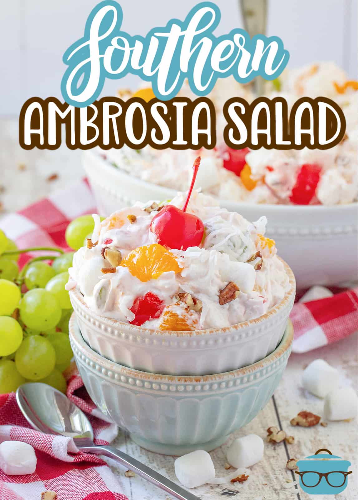 Southern Ambrosia Salad recipe from The Country Cook, a white and blue bowl stacked on top of each other, a serving of ambrosia salad shown in top bowl with a maraschino cherry on top and a bunch of green grapes in the background.