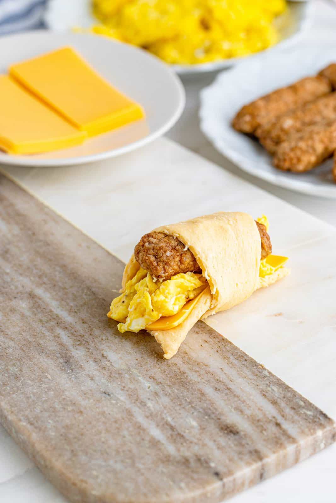 crescent roll shown wrapped around a sausage link, scrambled eggs and a slice of cheese. 