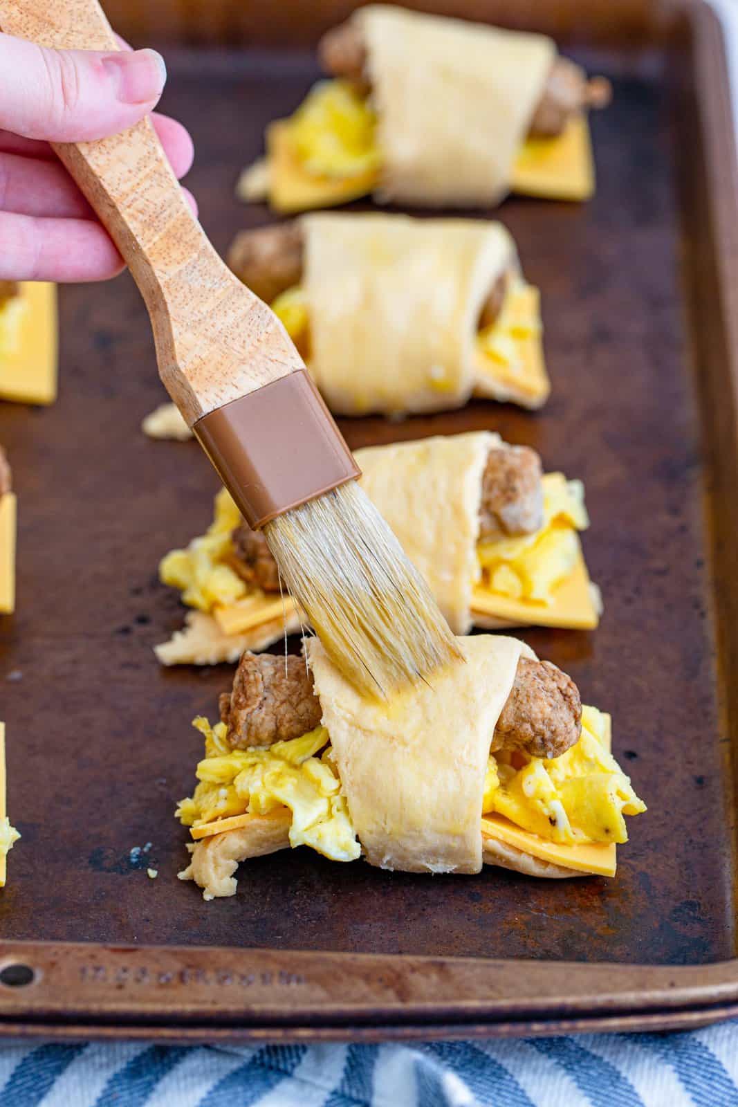 a pastry brush brushing a beaten egg on top of each crescent roll bundle. 