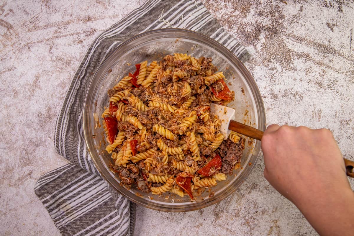 cooked rotini pasta, cooked ground beef and pizza sauce being stirred together in a large clear bowl