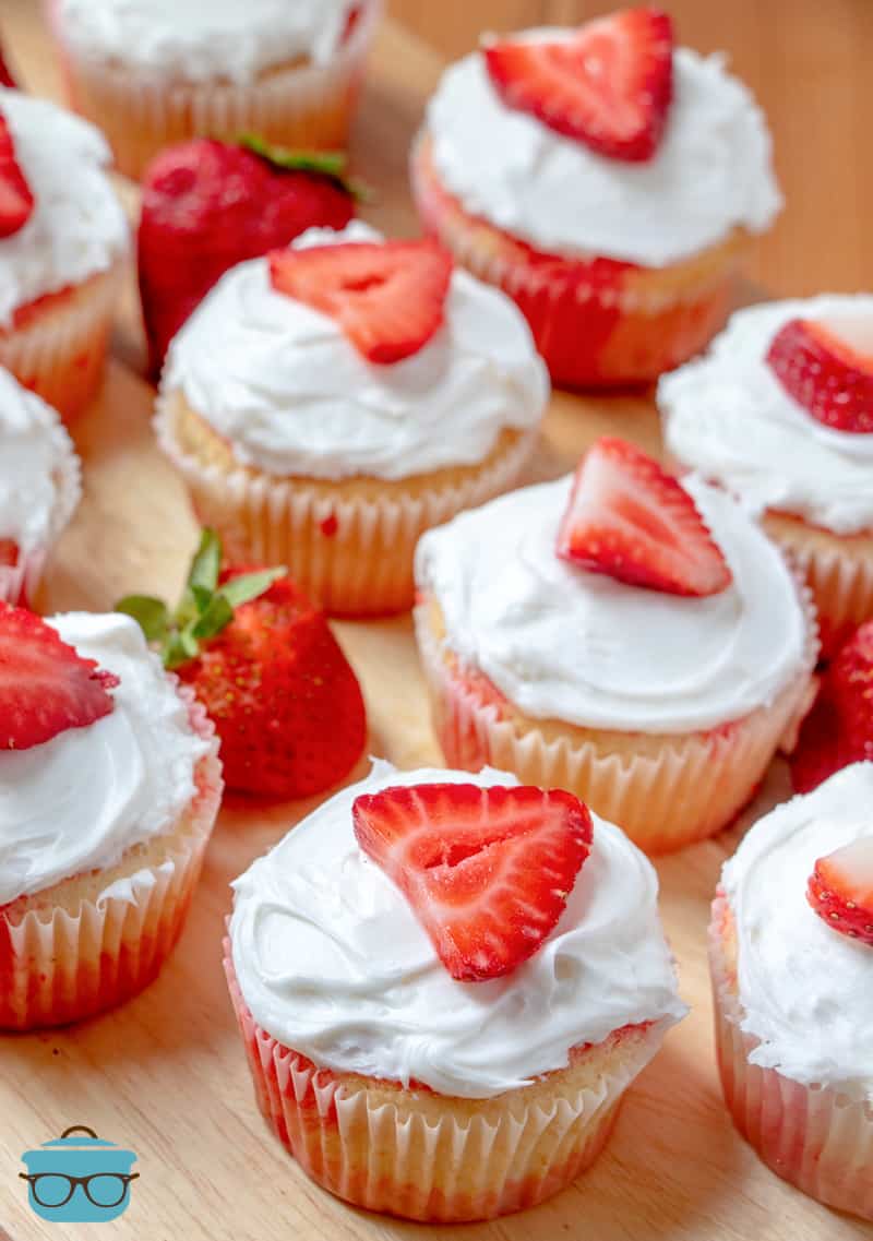 several frosted cupcakes topped a slices of fresh strawberries on a wooden cutting board. 