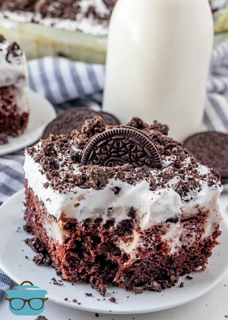 slice of Oreo cookie poke cake on a small white round plate with a milk bottle in the background, a bite has been removed from the cake.