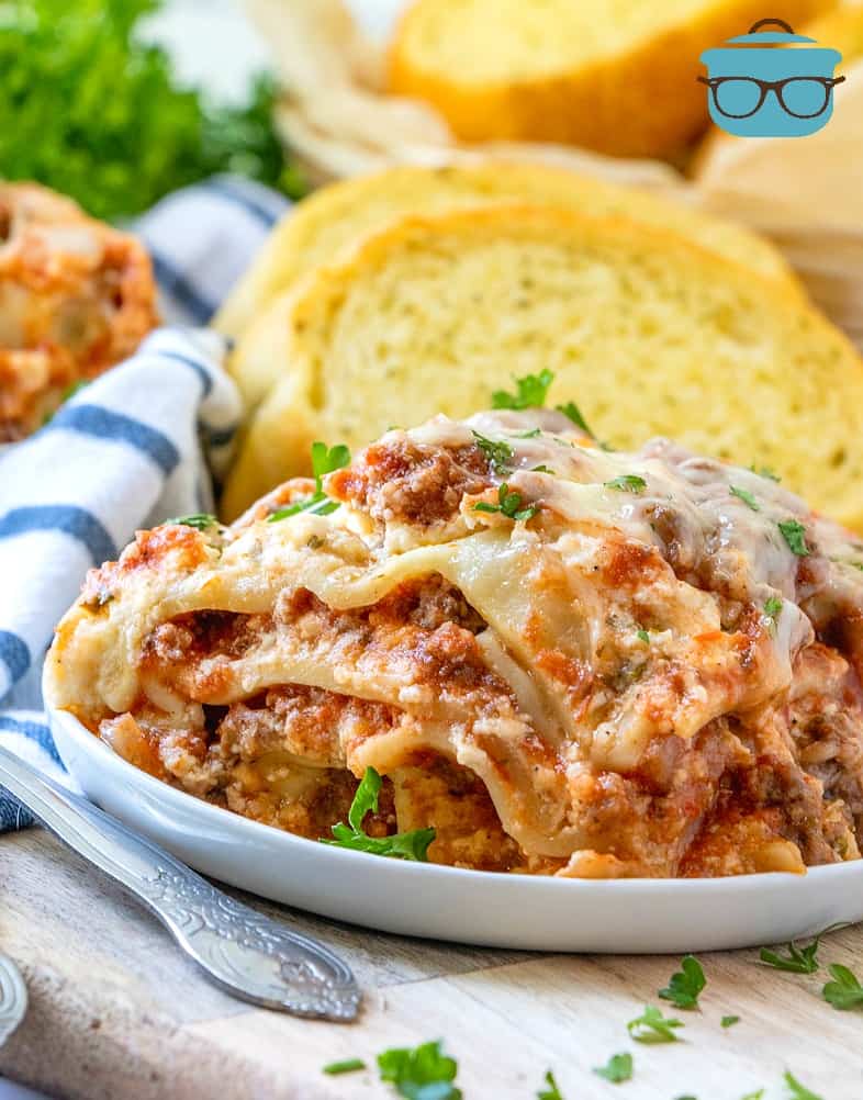 slice of slow cooker lasagna on a plate with a fork and slices of garlic bread.