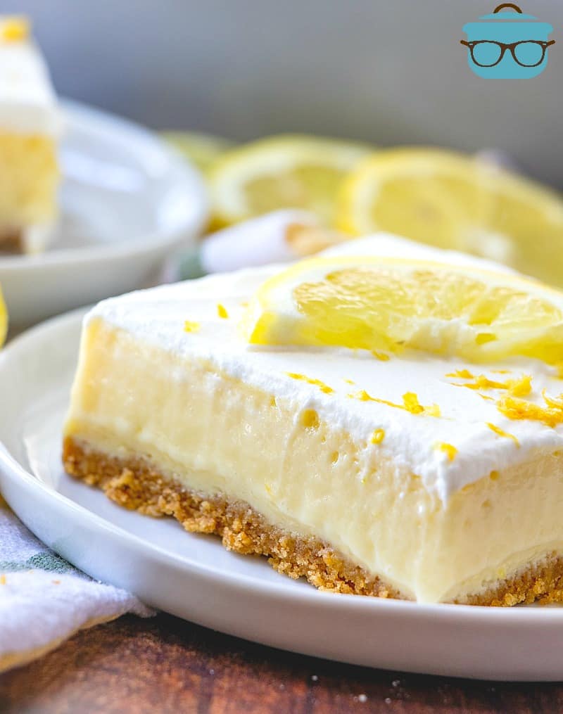 close up shot, slice of creamy lemon bar on a white plate, topped with a lemon slice and slices of lemon in the background.