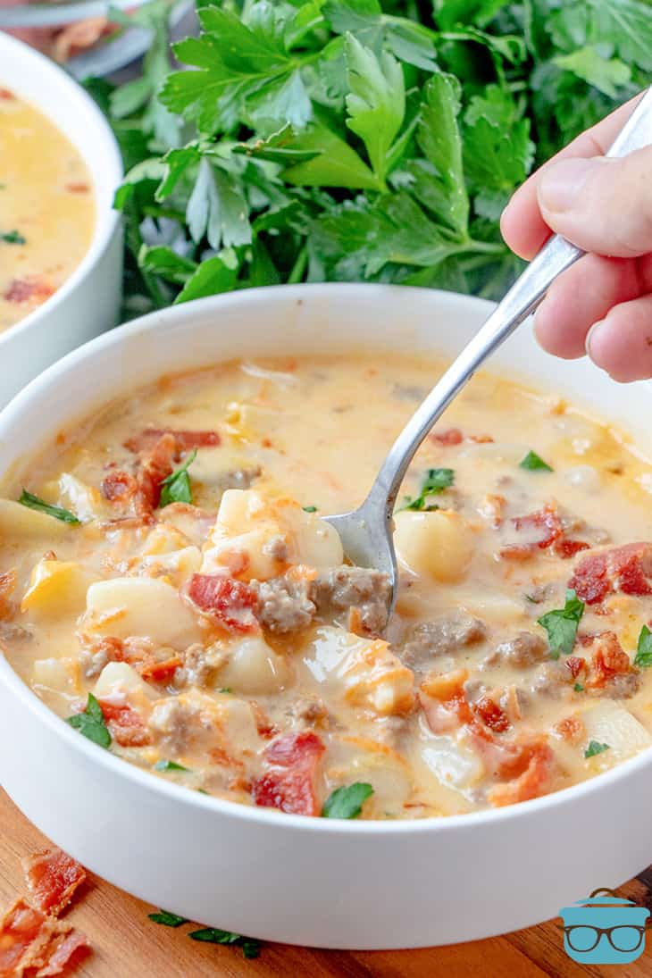 a spoon being inserted to a bowl of cheeseburger soup with a bunch of parsley in the background.