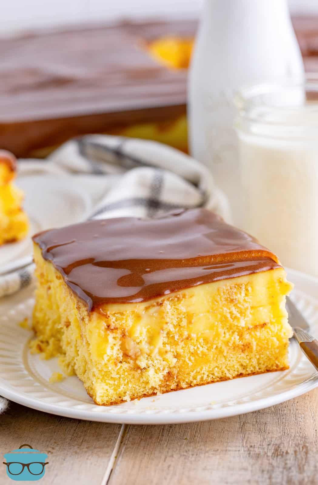 photo of Boston cream poke cake on a round white plate with a bottle of milk and a cake pan in the background. 