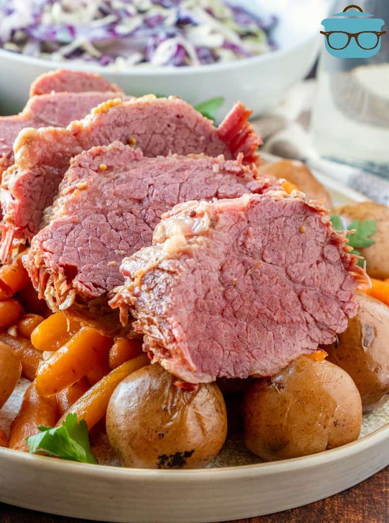 sliced corned beef on a bed of cooked potatoes and carrots with cole slaw in a bowl in the background.