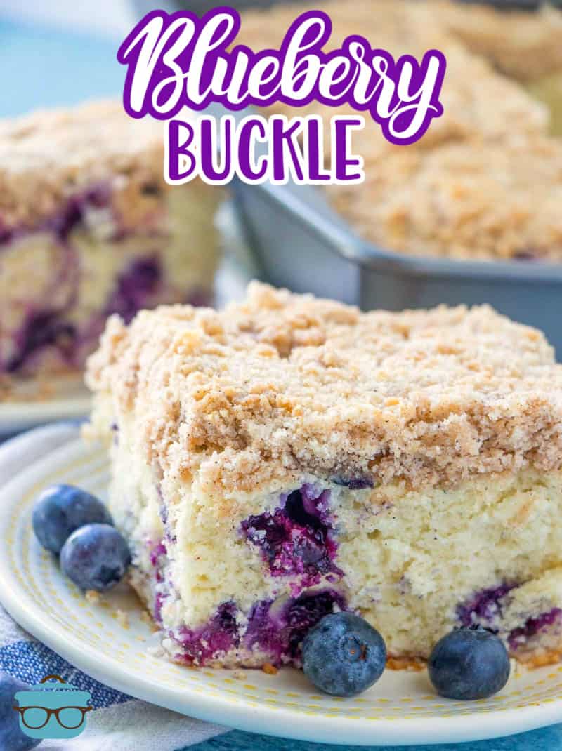slice of blueberry buckle cake on a small round white plate with fresh blueberries scattered around.