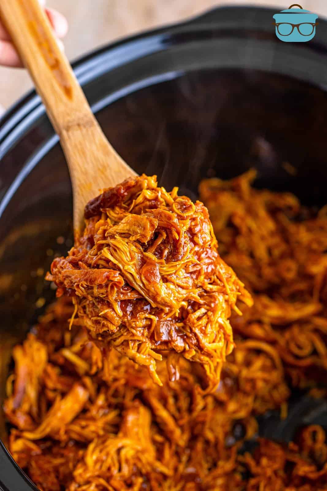 shredded BBQ chicken shown in a slow cooker with a wooden spoon holding up some of the cooked chicken. 