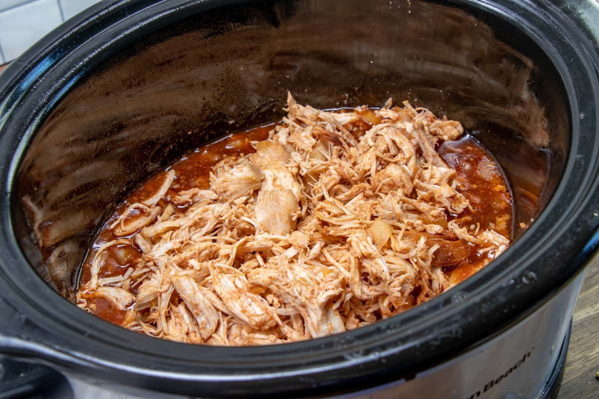 cooked, shredded chicken placed into a black oval slow cooker with barbecue sauce.