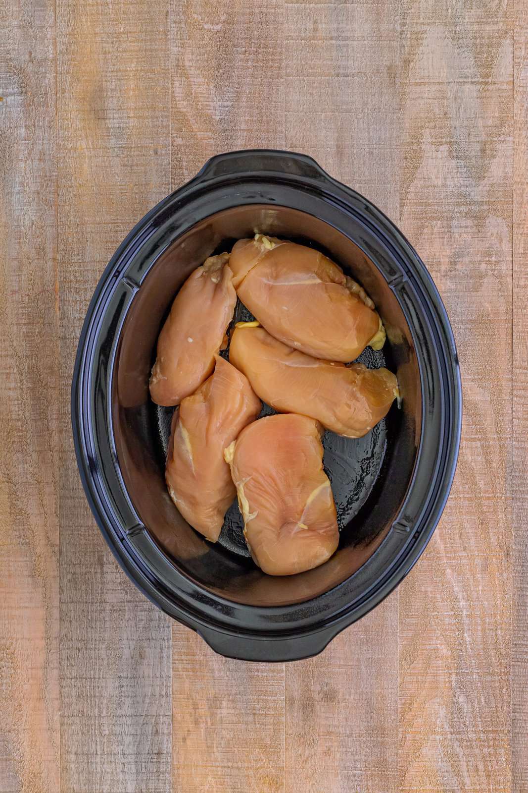 five raw chicken breasts shown in the bottom of an oval slow cooker. 