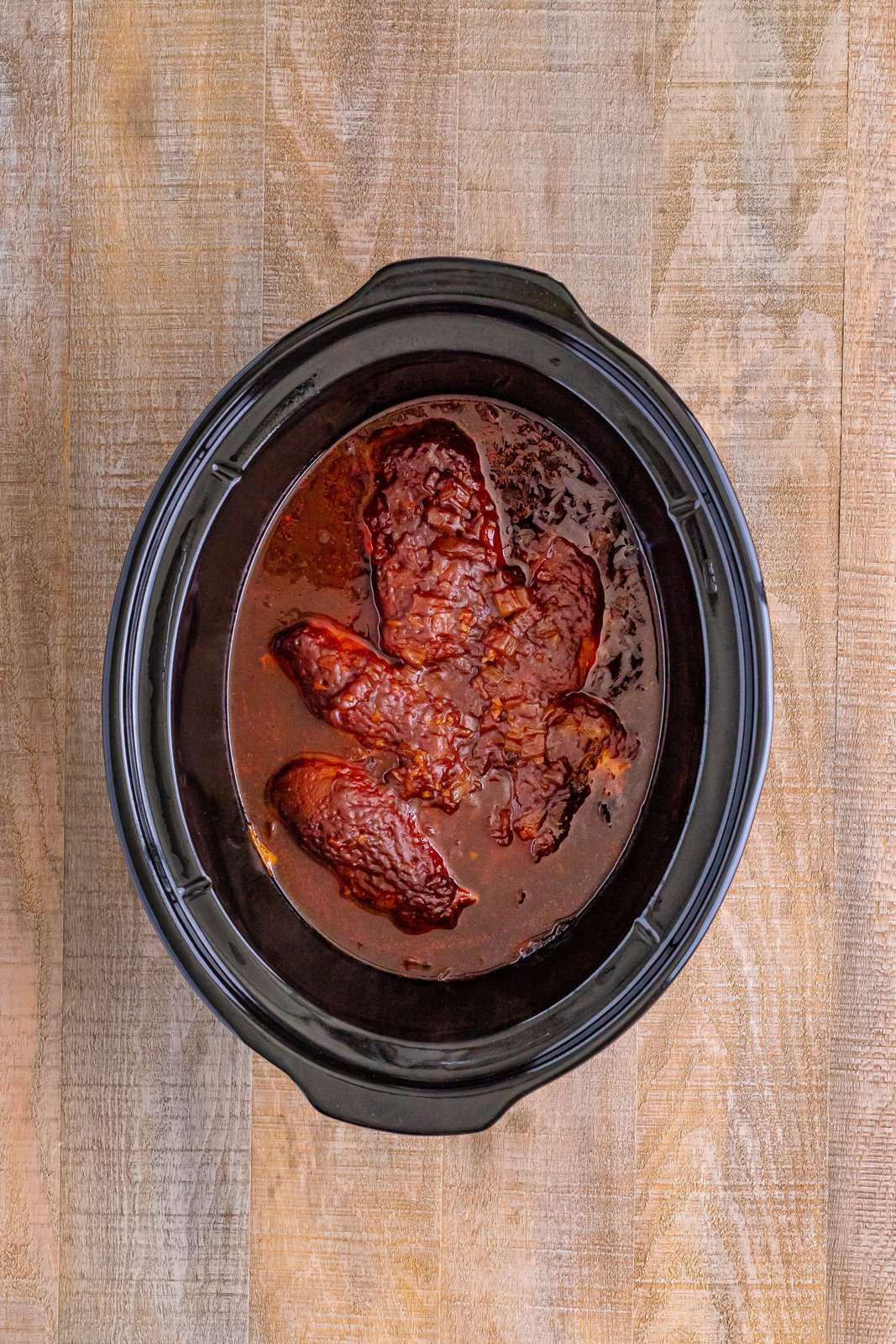 fully cooked chicken breasts in barbecue sauce shown in a black oval slow cooker. 