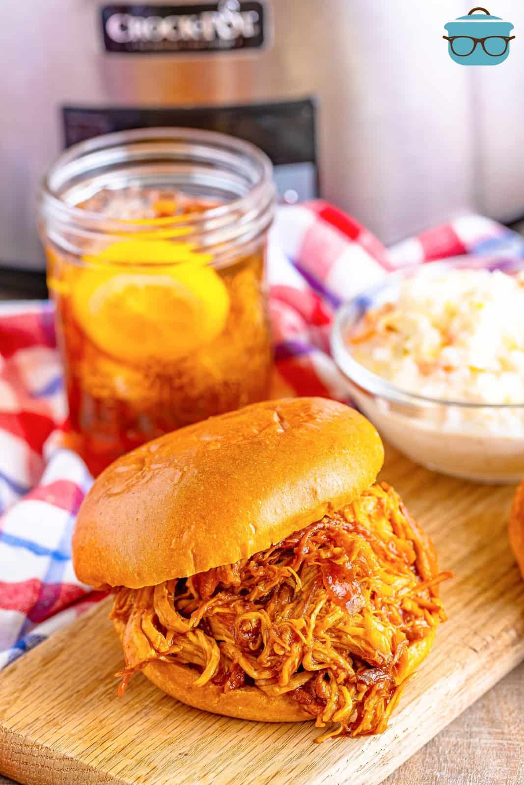 a shredded barbecue sandwich shown on a wooden cutting board with a small bowl of Cole slaw in the back and a mason jar of iced tea. 