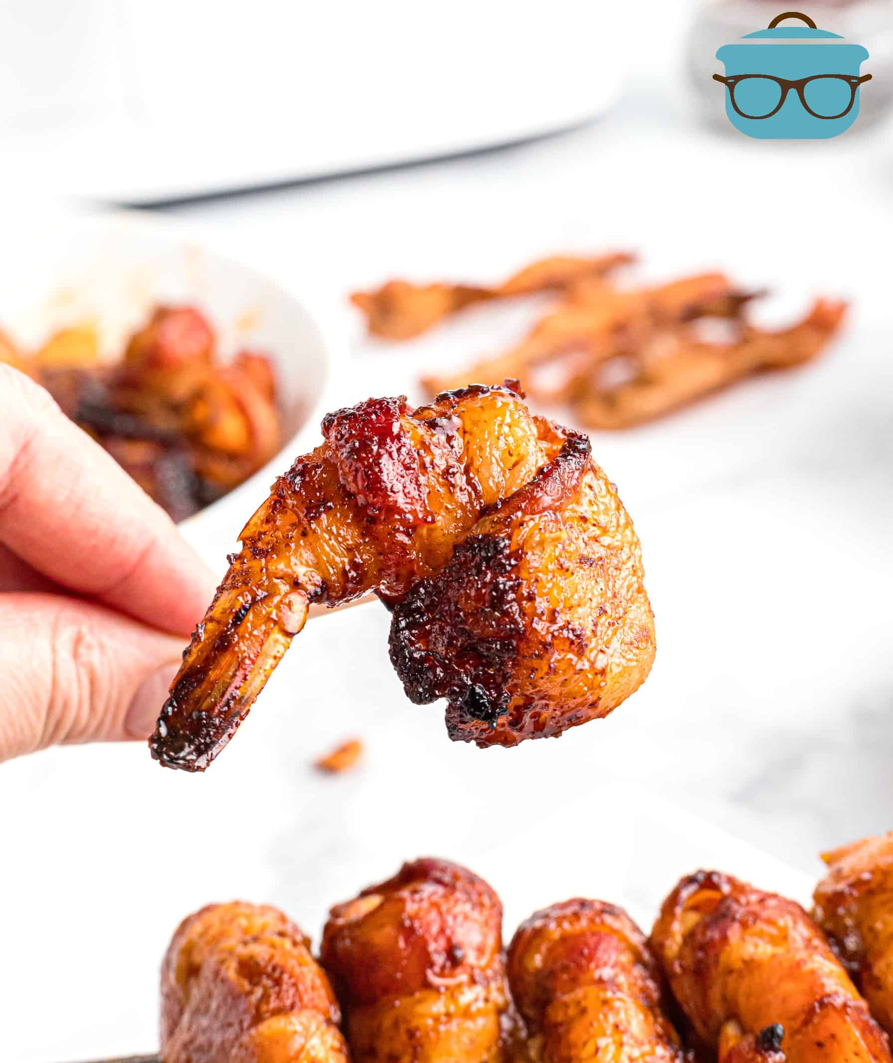 Bacon Wrapped Garlic Shrimp, close up picture of hand holding one bacon wrapped shrimp on a toothpick.