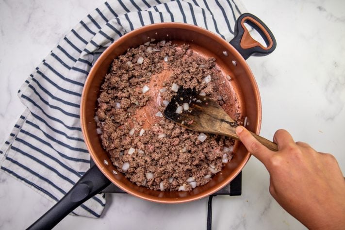 cooked ground beef, onions and garlic being stirred by a wooden spoon in a skillet.