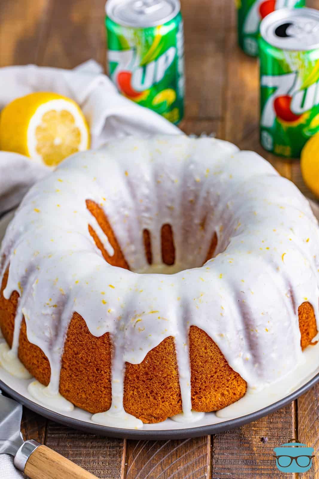 fully baked 7Up cake on a white cake plate with lemon icing drizzled on top and cans of 7Up shown in the background. 