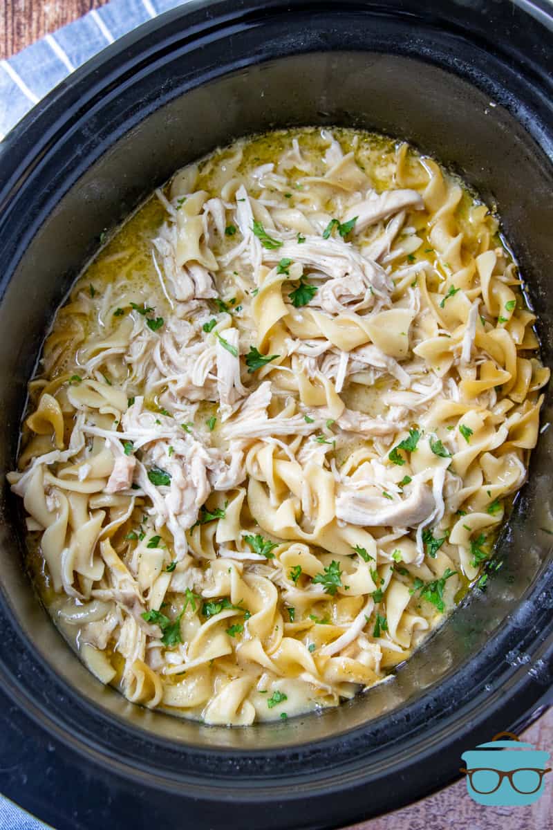 Fully cooked chicken and noodles in an oval black crock pot and topped with chopped parsley.