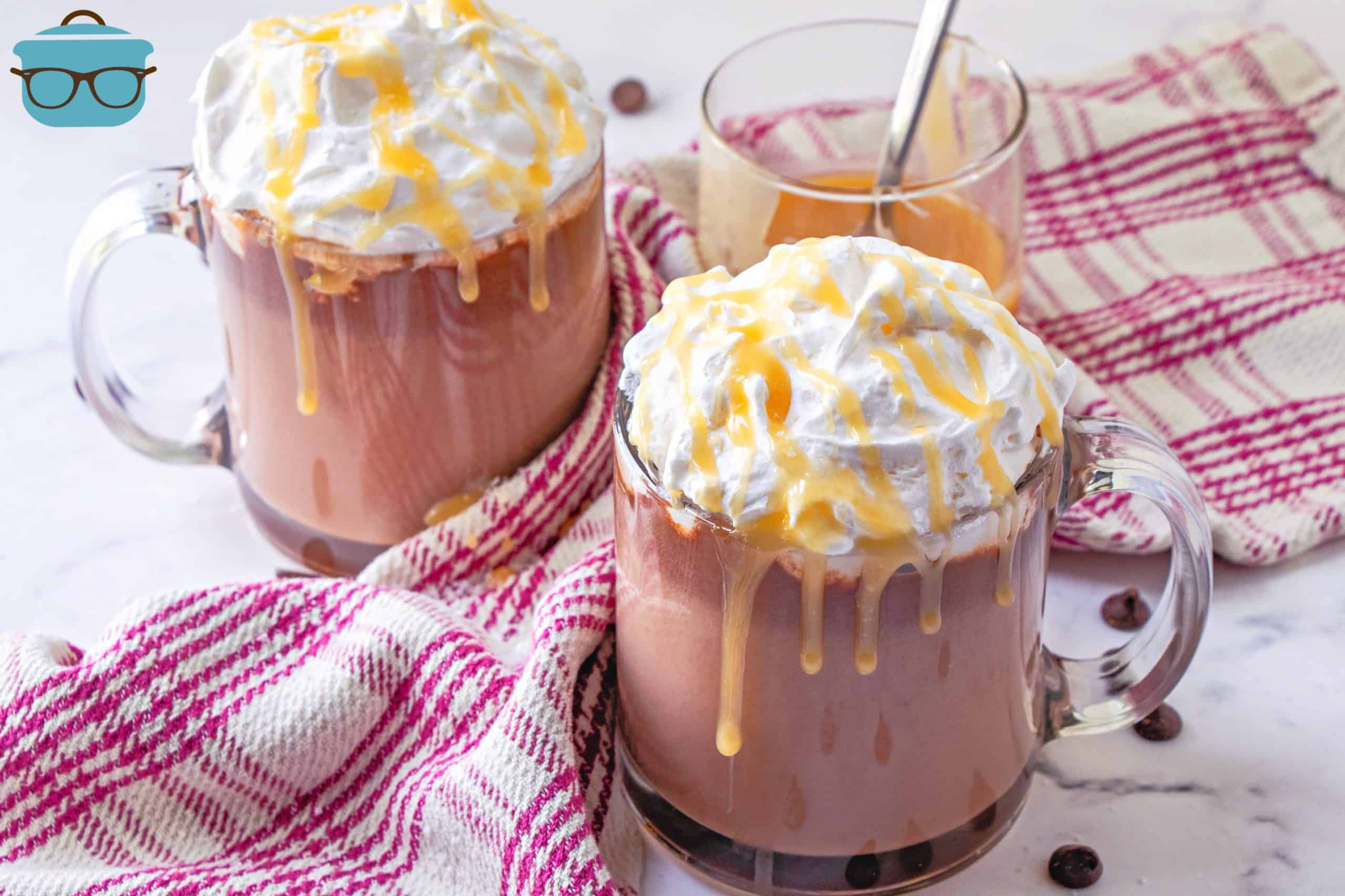 two cups of hot chocolate topped with whipped cream and drizzled with salted caramel sauce.