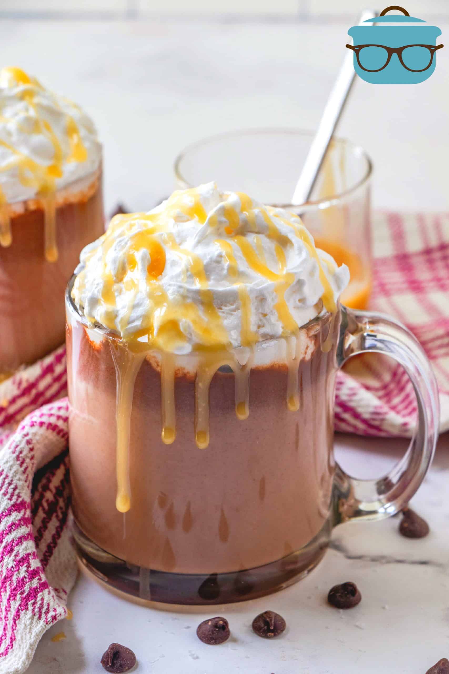 The Best Salted Caramel Hot Chocolate shown in a clear glass mug with chocolate chips scattered around and a red and white kitchen towel in the background.
