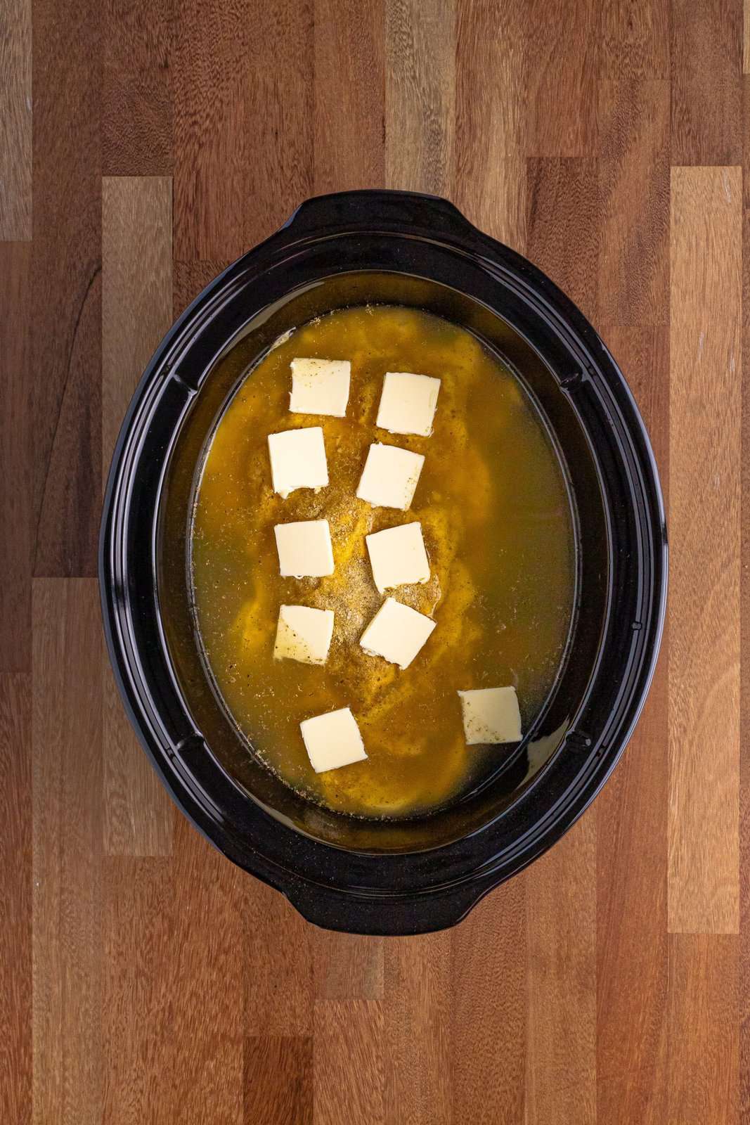 slices of butter shown with chicken and chicken broth in oval slow cooker. 