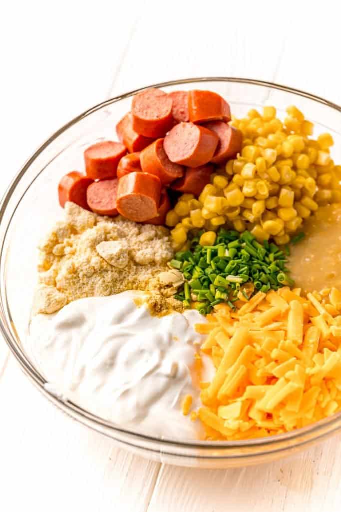 cream style sweet corn, whole kernel sweet corn, sour cream, salted butter, shredded cheddar cheese, Jiffy Corn Muffin Mix, minced chives, mustard powder, hot dogs in a large, clear mixing bowl