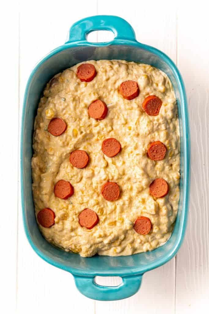 sliced hotdogs placed evenly around the top of the corn casserole batter in a blue baking dish