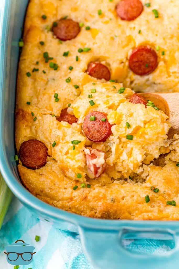 HOT DOG CORN CASSEROLE SHOWN IN A TEAL BAKING DISH WITH A WOODEN SPOON SCOOPING OUT A SERVING