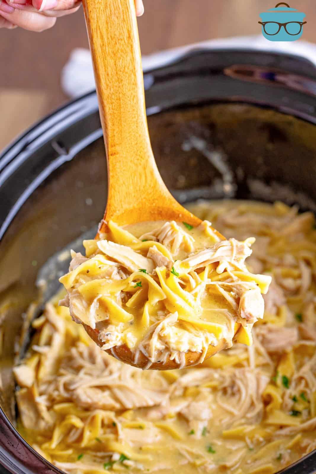 a wooden spoon scooping up some chicken and noodles out of the slow cooker. 