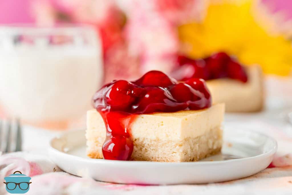 slice, sugar cookie cheesecake bars on a plate topped with cherry pie filling