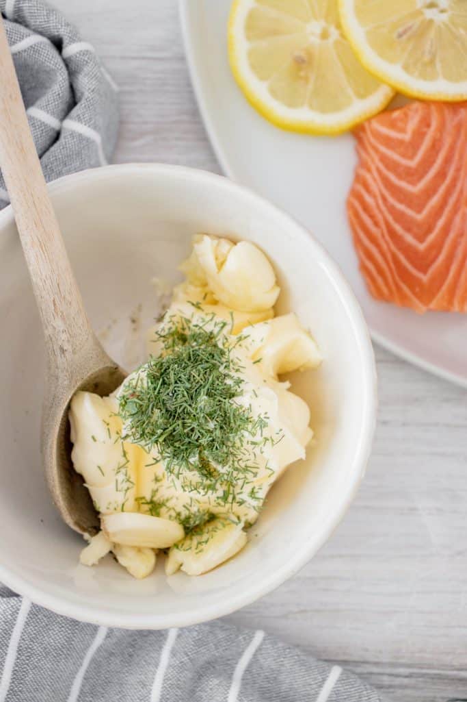 softened butter, garlic and dried dill in a white bowl and a wooden spoon