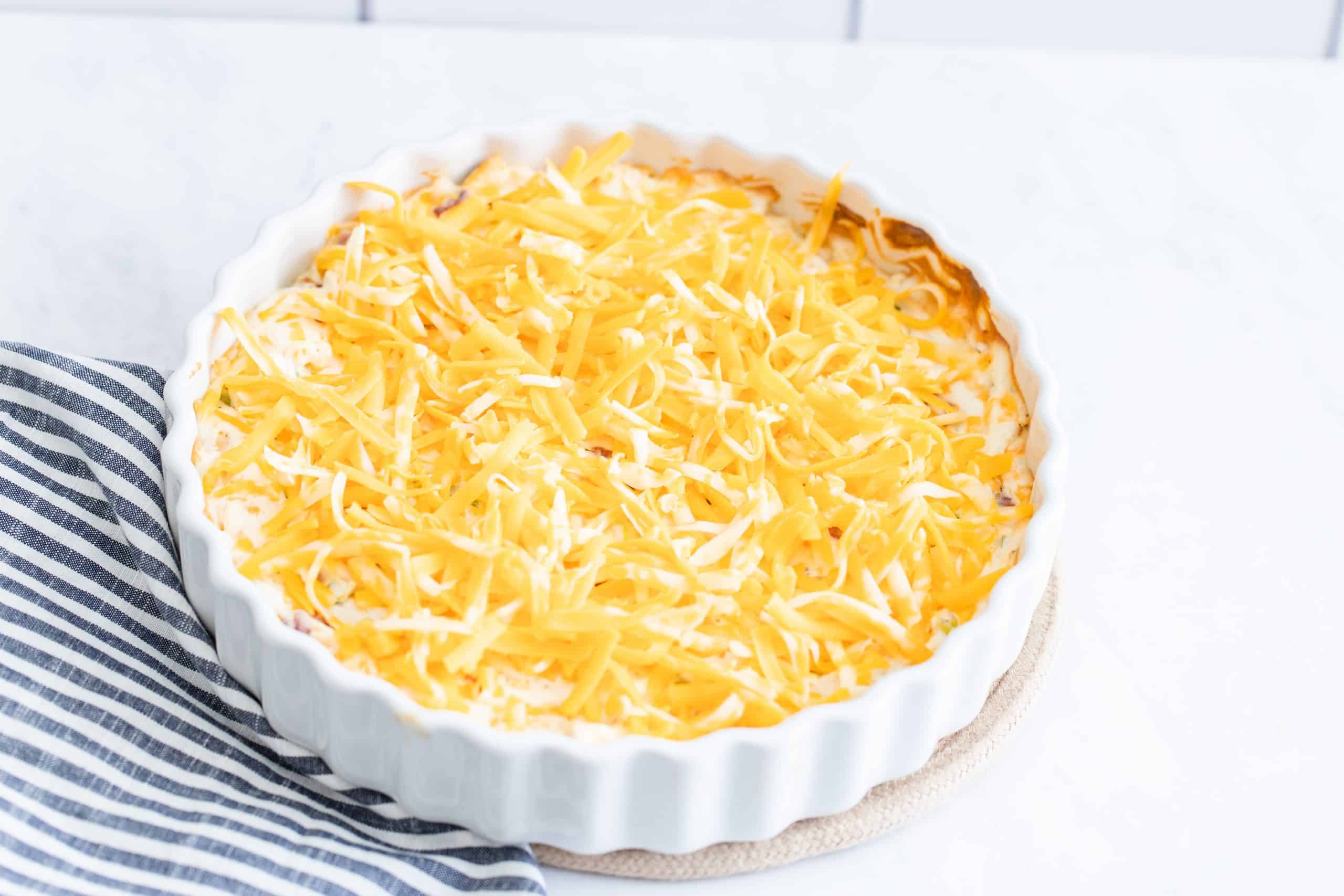 shredded cheese on top of baked cream cheese mixture in a white tart baking dish.