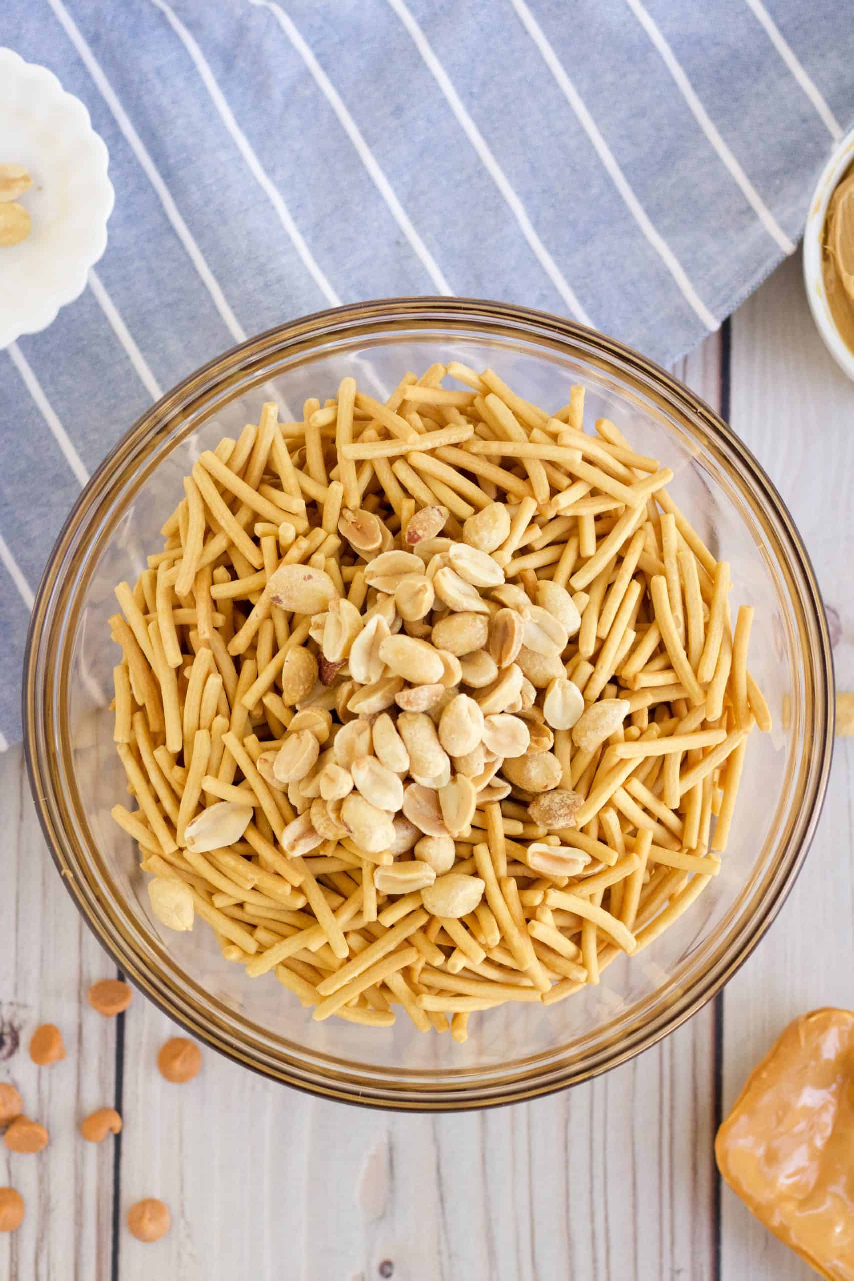 chow mein noodles and roasted peanuts added to bowl with melted butterscotch and peanut butter in a clear glass bowl.