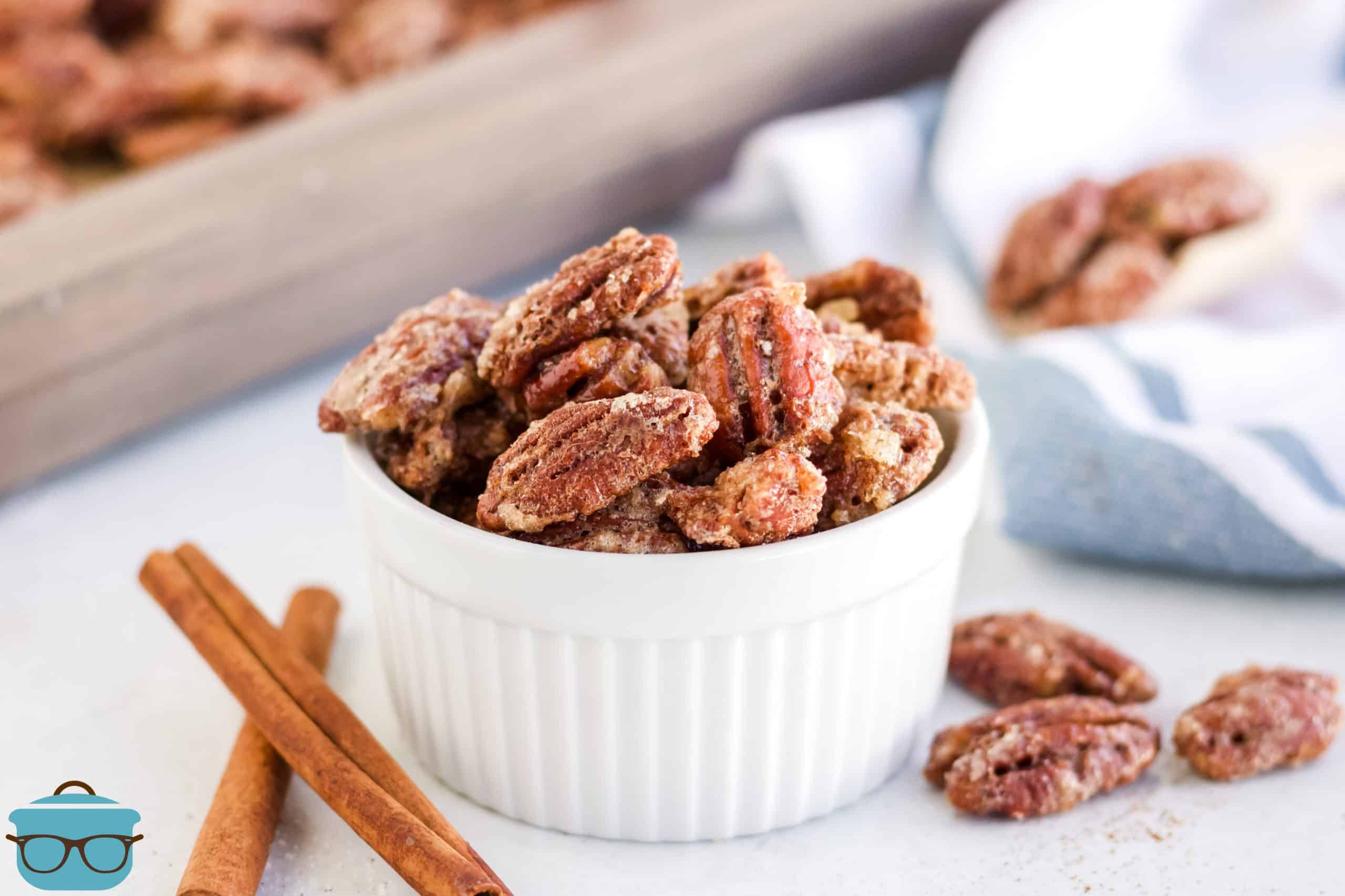 candied pecans shown in a small white ramekin bowl with cinnamon sticks on the side.