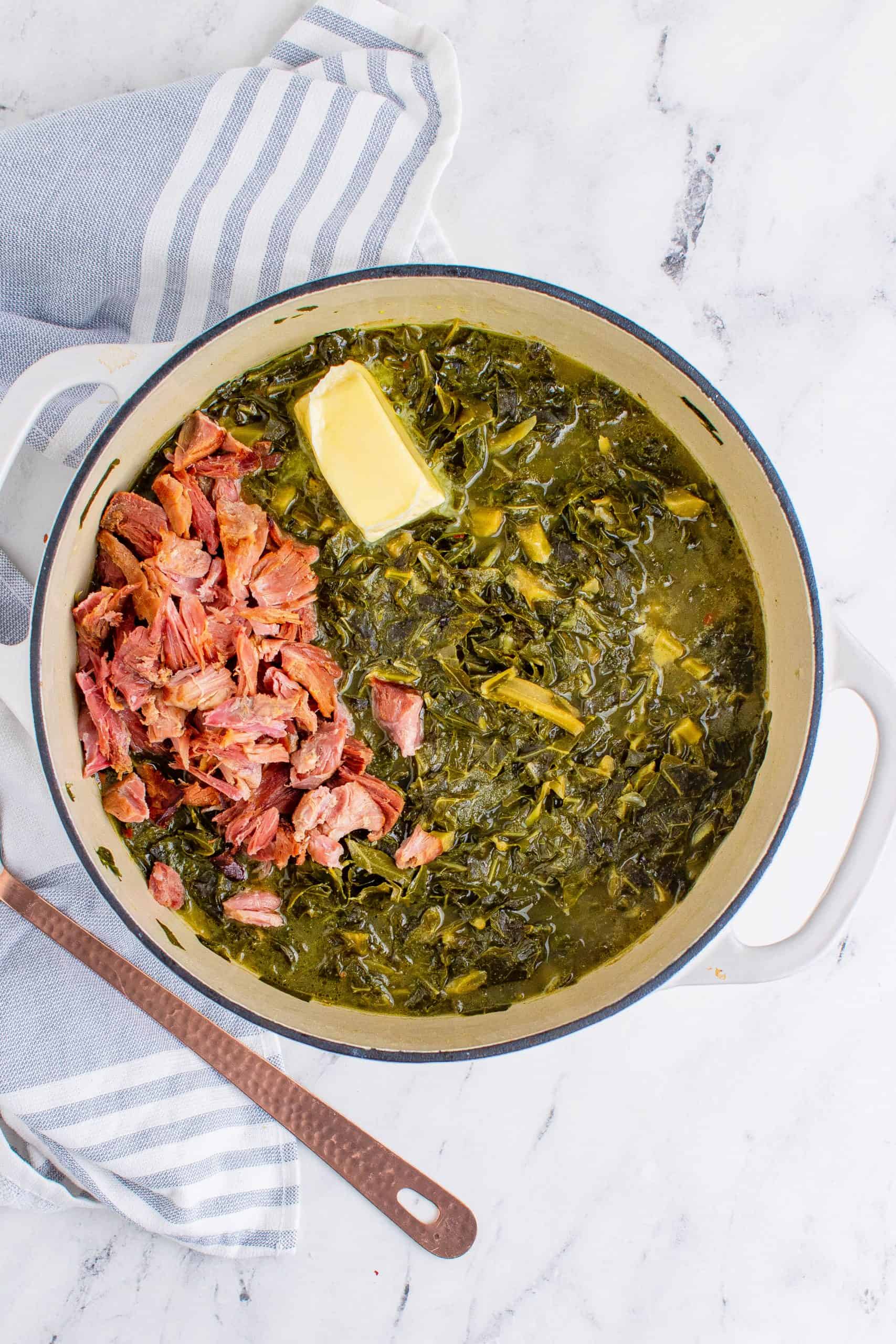 shredded ham added cooked collard greens and butter in a a white pot with a copper spoon on the side