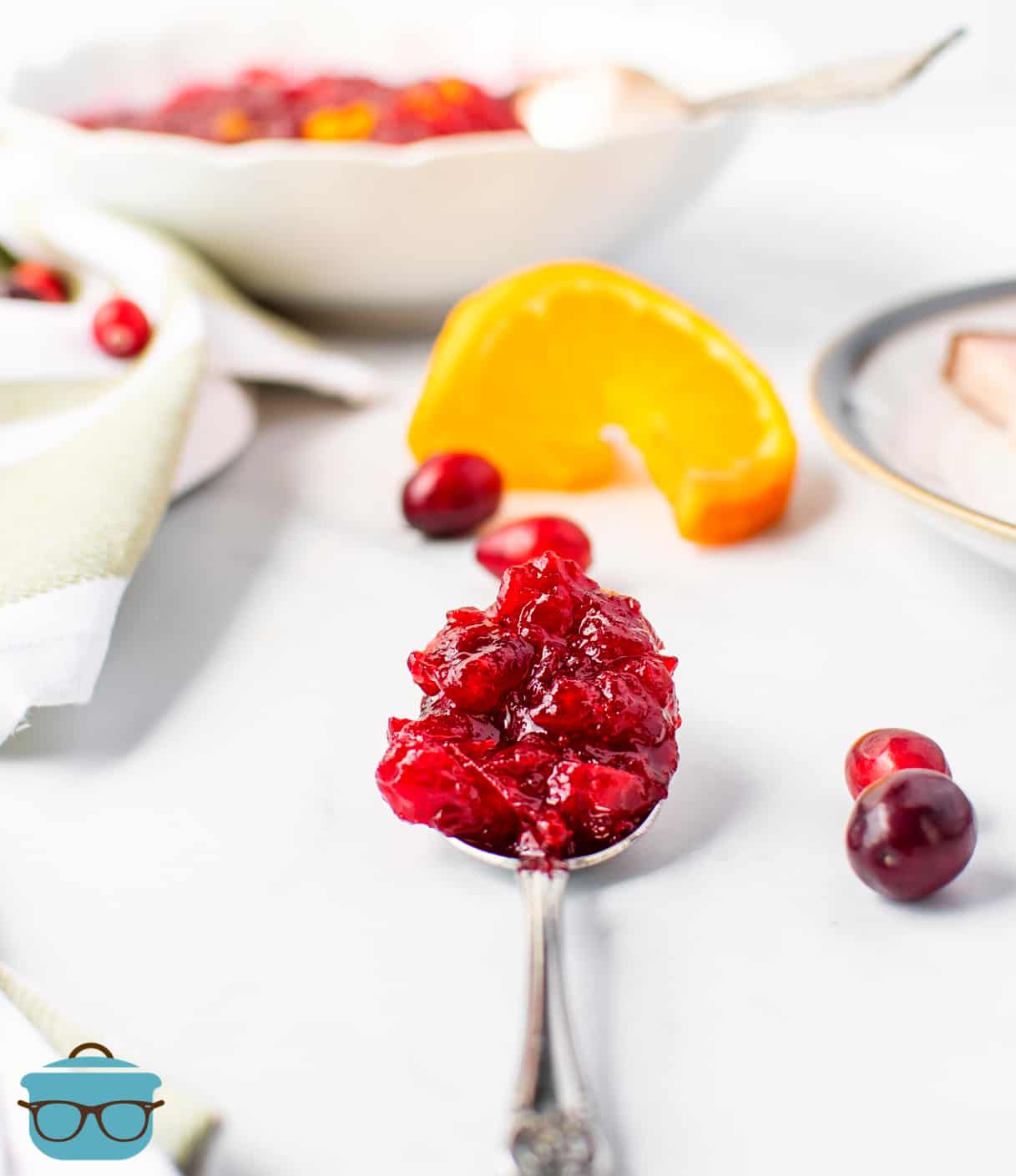 Cranberry sauce shown on a serving spoon on a white table with fresh cranberries scattered around.