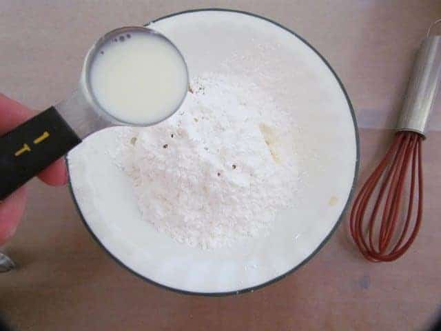 milk being poured into a white bowl containing powdered sugar and clear vanilla extract