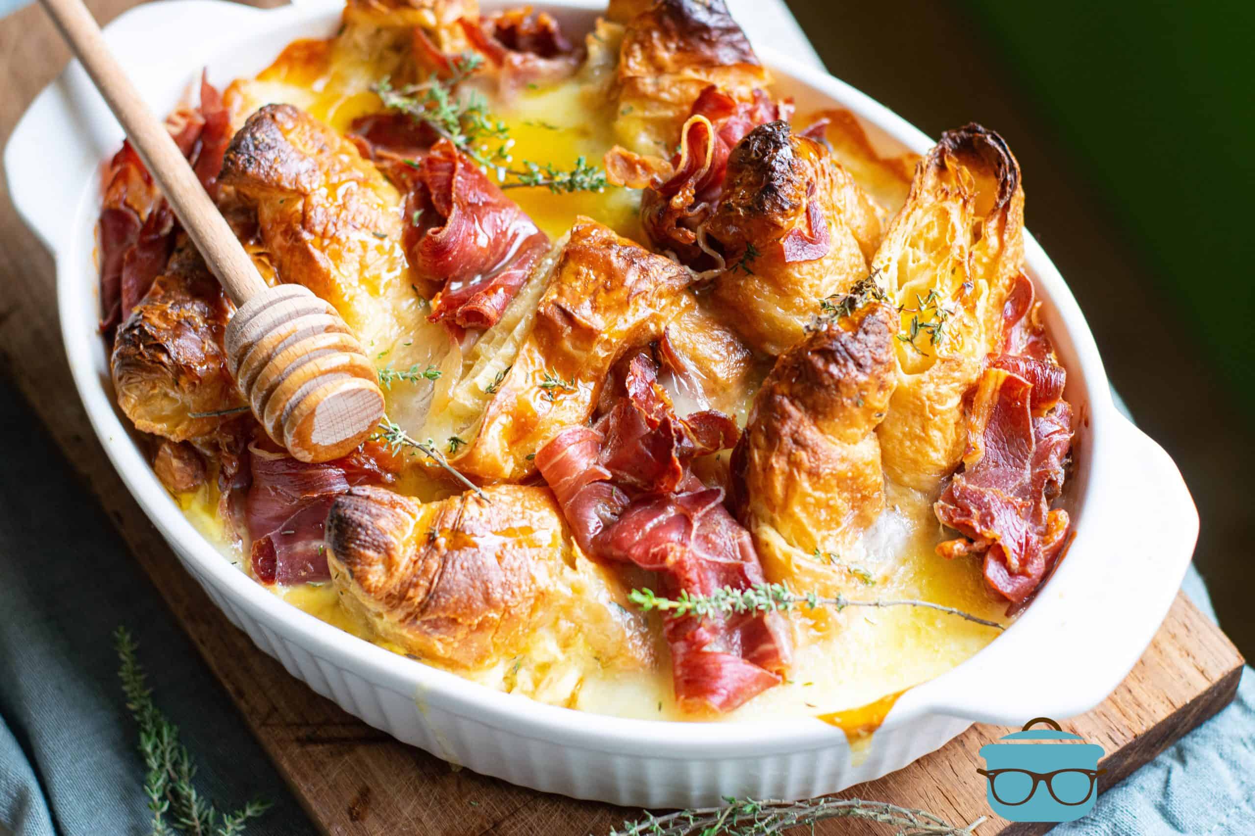 fully baked ham and cheese croissant bake in a white oval baking dish on top of a wooden cutting board.
