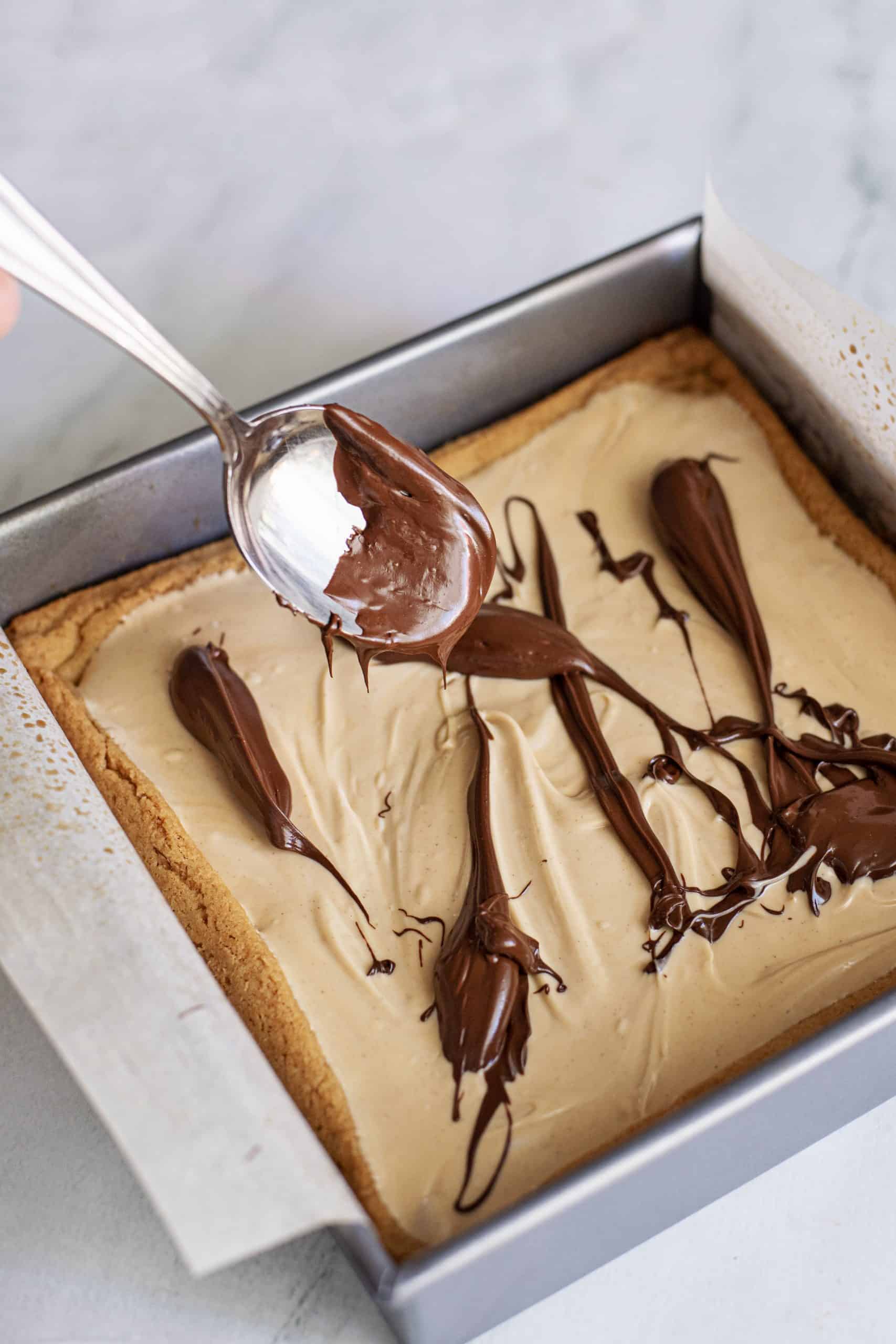 spooning melted chocolate over white cholate mixture on sugar cookie bars.